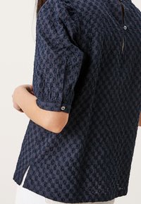 Blusa in blu navy con texture e motivo floreale in rilievo, maniche a sbuffo corte e chiusura con bottoni sul retro. Fessure laterali all'orlo.
