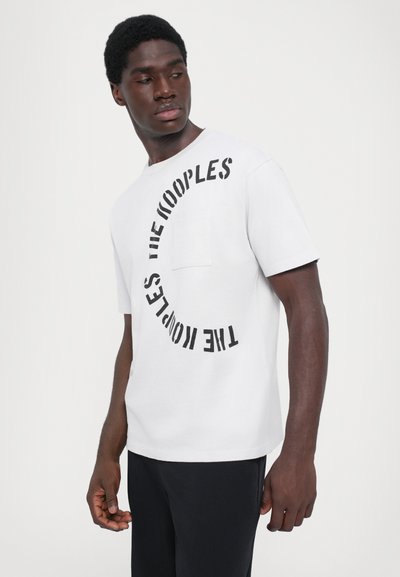 Biała bawełniana koszulka z okrągłym czarnym napisem "THE KOOPLES" oraz kieszonką na piersi. Krótkie rękawy i luźny krój.