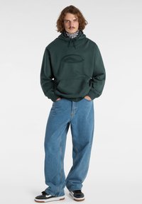 Felpa verde scuro con tasca frontale e materiale a loope, abbinata a jeans larghi in denim blu. Sneakers nere e bianche completano l'outfit.