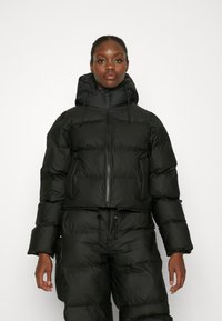 Svart kort pufferjacka med huva, dragkedjestängning, dragsnörningsdetaljer och puffy-textur; i kombination med matchande pufferbyxor.