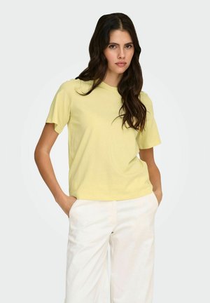 Jonge vrouw met lang donker haar, gekleed in een lichtgeel shirt met korte mouwen en witte broek, staand met handen in de zakken tegen een witte achtergrond.