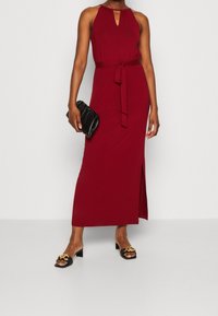 Robe maxi bordeaux avec un col halter, détail en trou de serrure, taille ceinturée et fente sur le côté, accompagnée d'une pochette noire et de sandales.