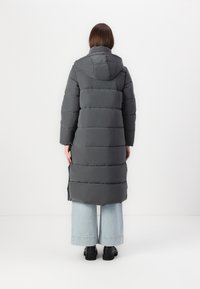 Lang grå pufferjakke med hætte, der har et quiltet design og sideslids, parret med lyseblå bredbenede jeans og sorte sko.