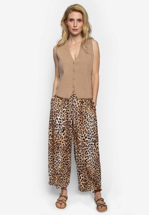 Vrouw met lang blond haar die een mouwloze bruine top met knopen draagt, brede broek met luipaardprint en bruine sandalen met bandjes, staand tegen een witte achtergrond.