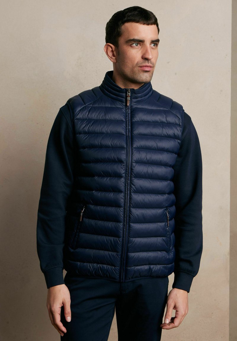Granatowa kamizelka puchowa z wysokim kołnierzem, poziomym pikowaniem, kieszeniami na zamek i gładką fakturą, noszona na ciemnej bluzie.