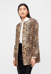 Σακάκι με λεοπάρ μοτίβο σε μπεζ και μαύρο, με στρογγυλή λαιμόκοψη, κουμπιά και μακριά μανίκια, συνδυασμένο με λευκό πουκάμισο.