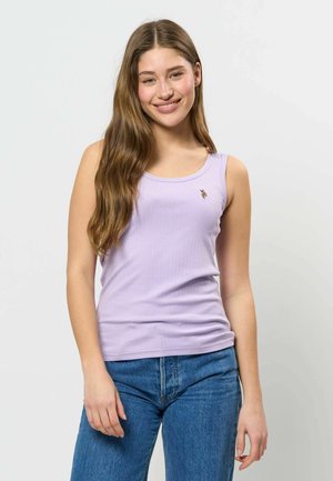 Ung kvinde med langt brunt hår iført en lyse lilla ærmeløs top og blå jeans, smilende mod en ensfarvet baggrund.