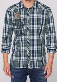 Man in een groen, marineblauw en witte geruite overhemd met opgestroopte mouwen en versleten blauwe jeans, zichtbaar van de taille tot de nek.