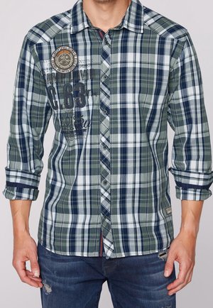 Man in een groen, marineblauw en witte geruite overhemd met opgestroopte mouwen en versleten blauwe jeans, zichtbaar van de taille tot de nek.