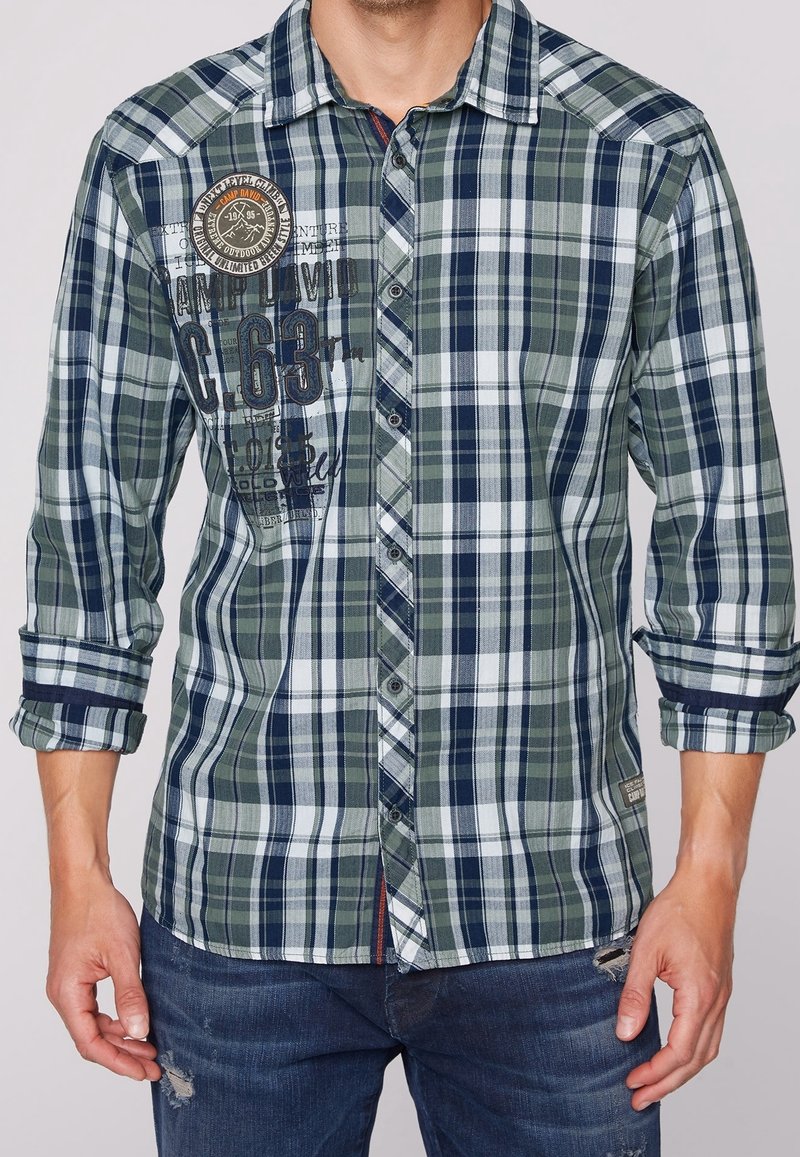 Man in een groen, marineblauw en witte geruite overhemd met opgestroopte mouwen en versleten blauwe jeans, zichtbaar van de taille tot de nek.