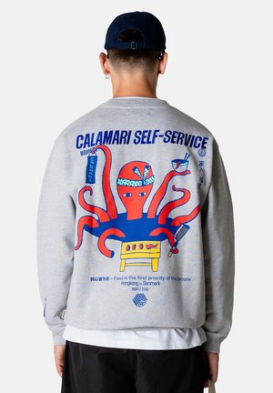 Person trägt grauen Pullover mit buntem Oktopus, der einen Grill, eine Schüssel mit Essen und ein Hackmesser hält, sowie den Schriftzug "Calamari Self-Service" auf dem Rücken.