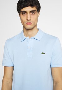 Polo shirt azzurro chiaro con una tessitura a trama, colletto con tre bottoni e logo di coccodrillo verde ricamato sul lato sinistro del petto. Maniche corte.