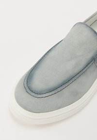 Chaussure slip-on en daim grise avec un bout rond, des accents brodés et une semelle en caoutchouc blanche. Texture lisse avec un effet dégradé.