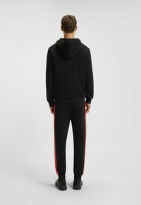 Felpa nera con cappuccio e maniche lunghe, abbinata a pantaloni da jogging neri con strisce rosse ai lati. Tessuto morbido, vestibilità atletica.