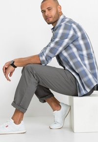 Camicia a quadri blu e grigia con maniche arrotolate, pantaloni grigi e sneakers bianche, seduto su un piedistallo bianco.