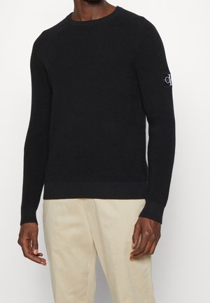 Mężczyzna ubrany w czarny, dzianinowy sweter z długim rękawem z naszywką z logo na lewej rękawie oraz jasnobeżowe spodnie, stojący na białym tle.