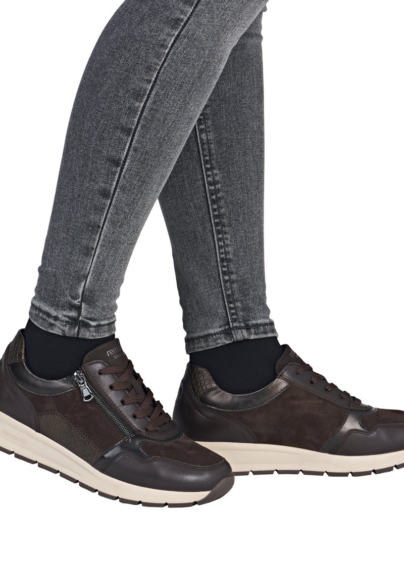 Braune Leder- und Wildledersneaker mit einer weißen Sohle, ausgestattet mit einem seitlichen Reißverschluss, Schnürsenkeln und strukturierten Akzenten. Getragen mit grauen Skinny Jeans.