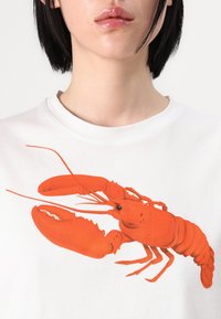 T-shirt branca com um grande gráfico detalhado de uma lagosta vermelha, exibindo suas garras e corpo segmentado impressos de forma intrincada no tecido.