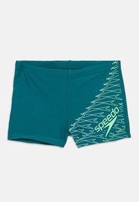 Žalio-žydros spalvos vyriški maudymosi šortai su ryškiai žaliais zigzagų raštais ir Speedo logotipu ant vienos pusės, išdėstyti plokščiai ant balto fono.