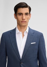 Blazer blu scuro in tessuto testurizzato, con rever a punta, chiusura a un bottone e un fazzoletto bianco nella tasca. Camicia bianca con colletto sotto.