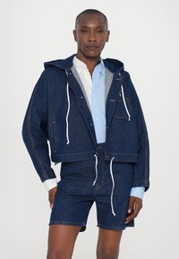 Denimjakke med hætte og frontlomme, med hvide snører og kontrasterende syninger, parret med matchende denimshorts.