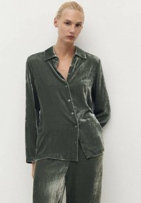 Camisa de terciopelo verde oliva con cuello y cierre de botones. Mangas largas con una textura suave y un brillo sutil.