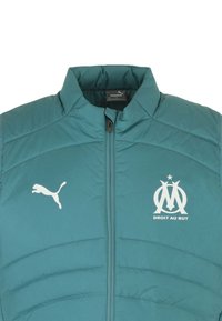 Giacca imbottita teal con colletto alto, zip frontale e logo Puma bianco. Sul petto è presente l'emblema "DROIT AU BUT" in bianco.