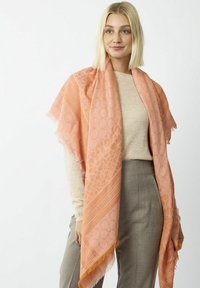 Helles Korallen-Schal mit geometrischen und Tiermustern, fransigen Kanten, weicher Textur, über den Schultern drapiert über einem beigen Oberteil und grauen Hosen.