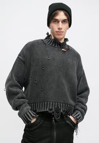 Suéter cinza curto com um colarinho franzido, detalhes desgastados e costura visível. Combinado com calças pretas, exibindo um tecido texturizado.