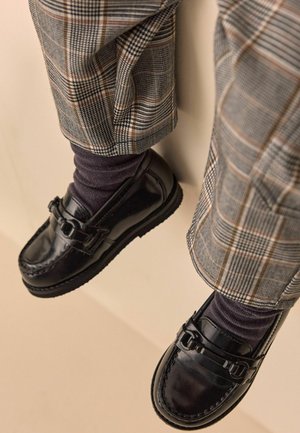 Mocassins en cuir noir avec une sangle et un détail de boucle, portés avec des chaussettes sombres et un pantalon à carreaux gris, mettant en avant une texture lisse et un design classique.