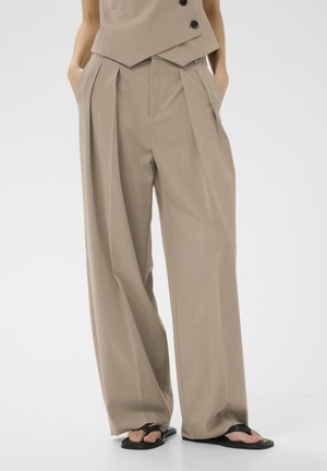 Beige high-waist wide-leg trousers paired with a matching vest, worn with black flip-flops, hands casually in pockets.