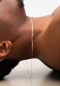 Goldene Kettenhalskette liegt auf der Haut und besitzt ein flaches, schlankes Design mit einer strukturierten Oberfläche und einem leichten Glanz auf dem Metall.