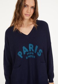 Navy blue knit sweater with a V-neck, featuring the text "PARIS MON AMOUR" in glittery blue. Includes two front pockets. Soft texture.