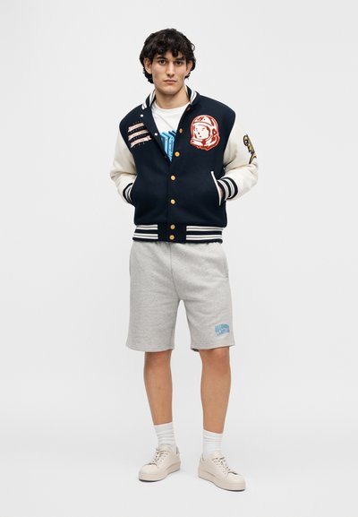 Ναυτικό μπλε varsity μπουφάν με κρεμ μανίκια, χρυσά κουμπιά και μπαλώματα. Γκρι βερμούδα με μπλε λογότυπο. Παπούτσια σε ουδέτερο τόνο, σύνολο σε απλό φόντο.