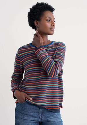 Frau mit kurzen lockigen Haaren, die ein bunt gestreiftes Langarmshirt und blaue Jeans trägt, schaut zur Seite und hat eine Hand an ihrem Hals.