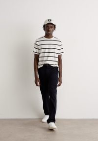 Hvidstribet t-shirt, sorte bukser og hvide sneakers. Modellen bærer en lys beige kasket med et bogstav "G." Enkel, afslappet outfit.