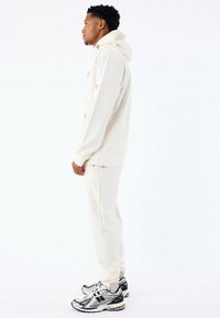 Sudadera beige con un bolsillo canguro, combinada con pantalones ajustados a juego y zapatillas deportivas en blanco y negro con toques de color vibrantes.