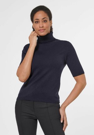 Femme portant un pull à col roulé manches courtes bleu marine et un pantalon noir, posant avec une main près du menton et l'autre sur la hanche.