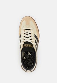 Beige lenkkari mustilla raidoilla, valmistettu mokasta ja nahasta, pyöreä kärki, teksturoitu pohja ja kultainen logo kielekkeellä.