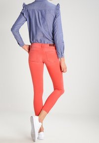 Mujer llevando jeans ajustados color coral, camisa de manga larga azul con hombros acampanados y zapatillas blancas, de pie con una pierna doblada.