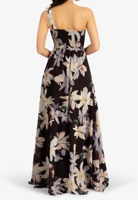 Femme portant une robe longue noire à une épaule, imprimée de grandes fleurs blanches et pastel, vue de dos avec les mains sur les hanches.