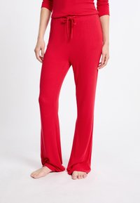 Rode geribbelde loungebroek met een verstelbare tailleband, wijde pijpen en een zachte stoffen textuur. Het model staat op blote voeten, waardoor de lengte van de broek goed zichtbaar is.