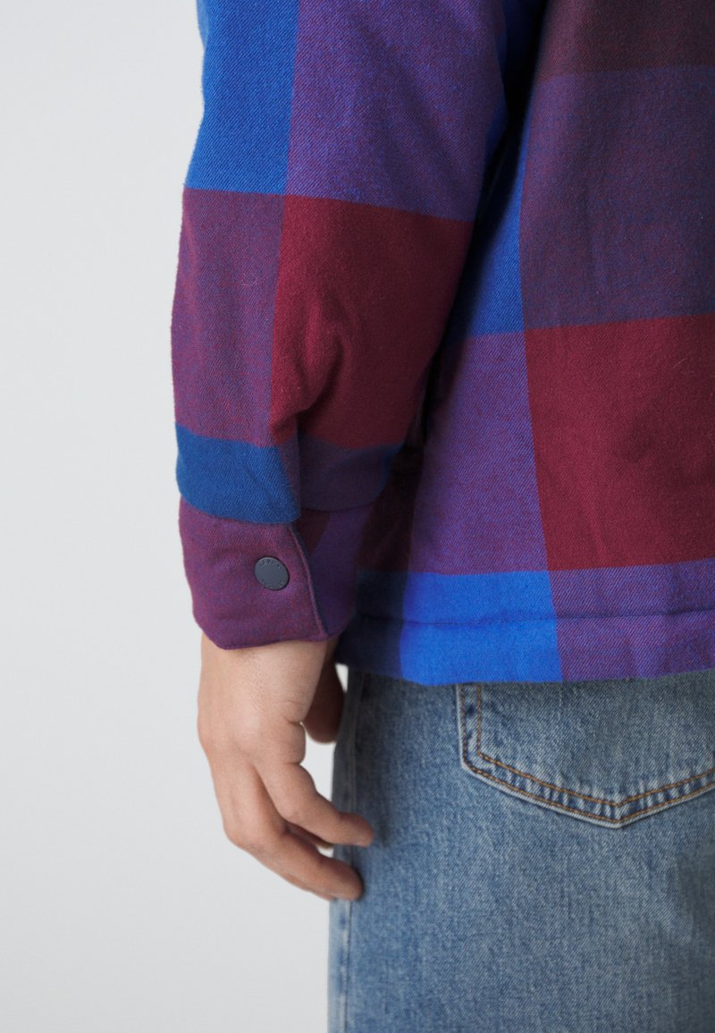 Flanellhemd mit einem blauen und burgunderfarbenen Karomuster, weicher Textur, Knopfmanschetten und lässiger Passform. Jeans sind in der Taille sichtbar.