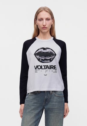 Femme aux cheveux blonds et lisses portant un t-shirt noir et blanc à manches longues avec un motif lèvres et le texte Voltaire, associée à un jean bleu délavé.