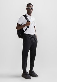Hvit polo skjorte med svarte detaljer, svarte joggebukser og svarte joggesko. Modellen bærer en svart ryggsekk og har på seg reflekterende solbriller.