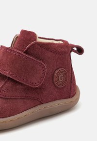 Burgundowe zamszowe buty z rzepem, okrągłym kształtem i teksturowaną gumową podeszwą. Posiadają okrągłe logo po zewnętrznej stronie.