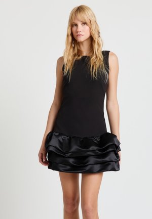 Donna con capelli biondi lunghi che indossa un abito nero senza maniche con gonna a balze di raso, in piedi davanti a uno sfondo grigio chiaro uniforme.