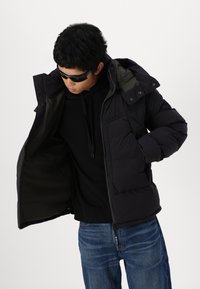 Svart vadderad jacka med avtagbar huva, frampockets och dragkedjestängning, bärs över en svart hoodie och blå jeans.