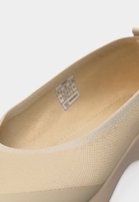 Μπεζ πλεκτό παπούτσι slip-on με στρογγυλεμένη μύτη και χαμηλό σφήνωτο τακούνι, που δείχνει λεπτομερή υφή και εσωτερική ετικέτα.