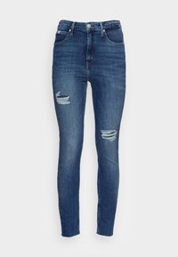 Jeans ajustados en denim azul oscuro, con áreas desgastadas en las rodillas, diseño de cinco bolsillos y una cinturilla estándar con cierre de botón y cremallera.